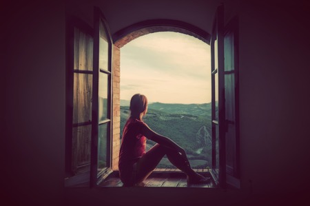 47060552 - young woman sitting in an open old window looking on the landscape of tuscany, italy. conceptual romantic, dreaming, hope, travel.